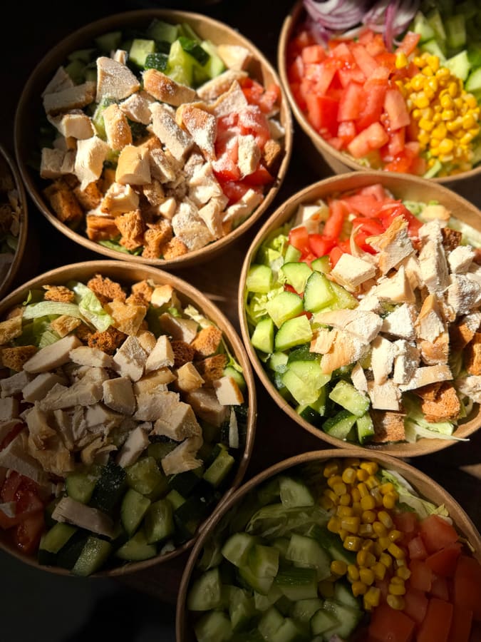 Five salad bowls in kraft containers seen from above, filled with iceberg, cucumber, tomato, corn, red onion, croutons and sliced chicken breast.