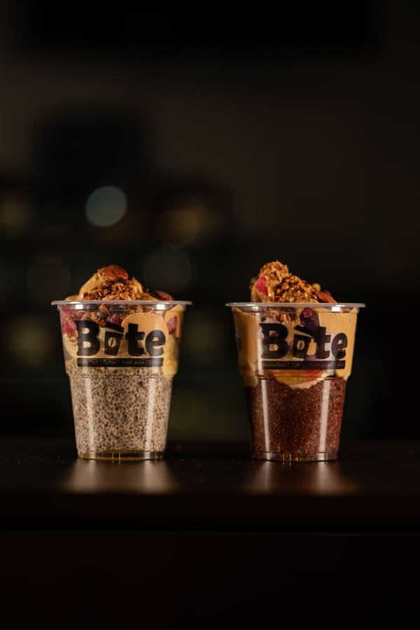 Two BITE chia pudding cups side by side, one plain and one with cocoa, both topped with granola, peanut butter, almonds and dried berries.