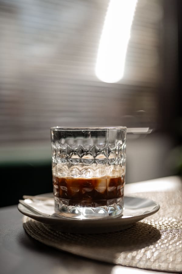 An iced coffee in a cut-glass tumbler on a saucer, backlit by afternoon light, with the milk and coffee still separated in layers.
