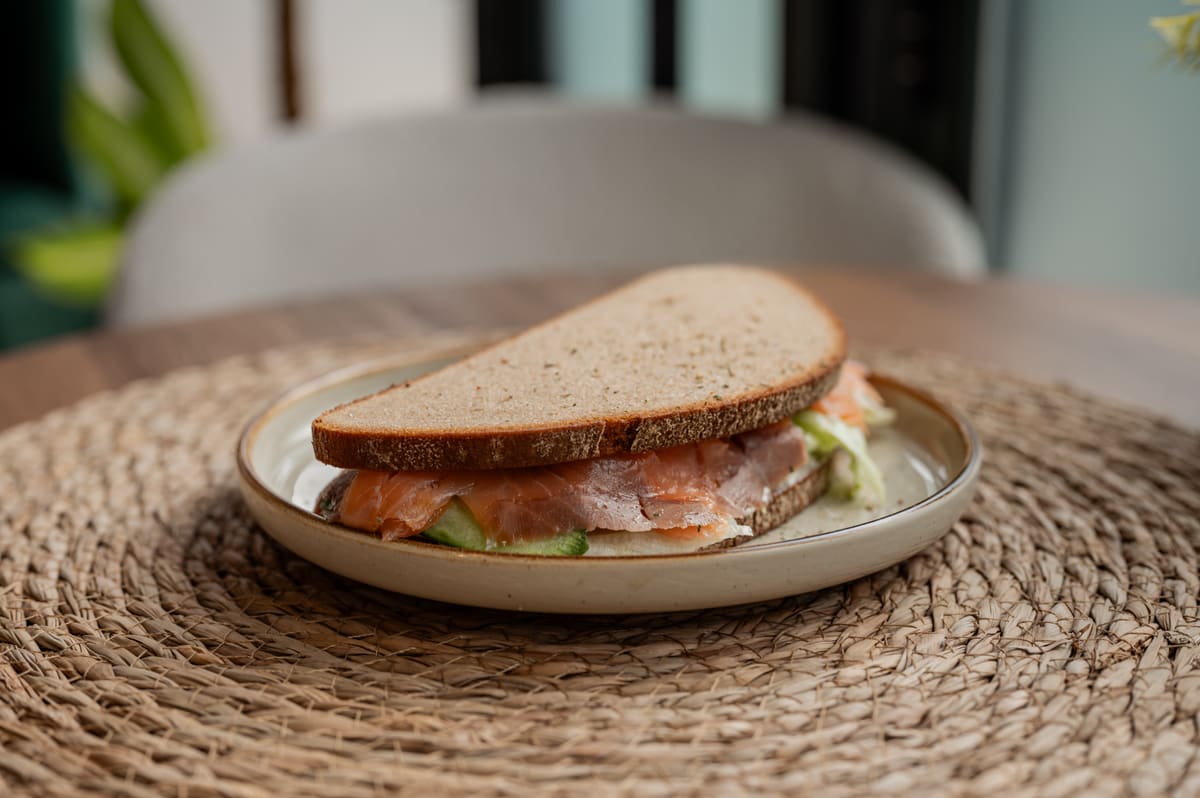 A smoked salmon sandwich on dark seeded bread with cucumber and lettuce, served on a ceramic plate on a woven placemat.