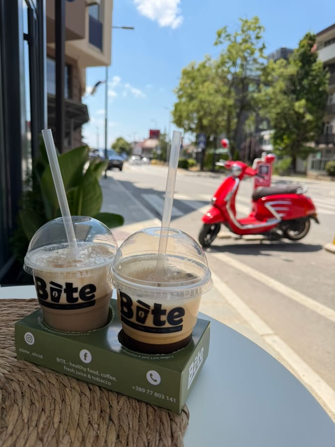 Two iced coffees in BITE cups on a takeaway tray at a terrace table, with Bulevar Makedonski Prosvetiteli and a parked red scooter behind.